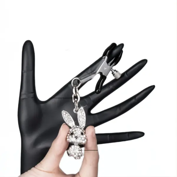 Close-up of a hand holding a silver nipple clamp with a dangling crystal bunny charm, clipped onto a black display hand.