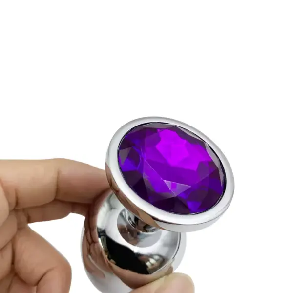 Close-up of a hand holding the large silver butt plug topped with an oval purple faceted gem, showcasing its reflective finish and scale against a white background.