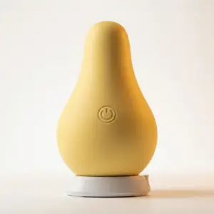 Pear Clit Vibrator – FruGasm in matte yellow silicone with a soft, rounded form and a central power button. Displayed upright on its white charging base.