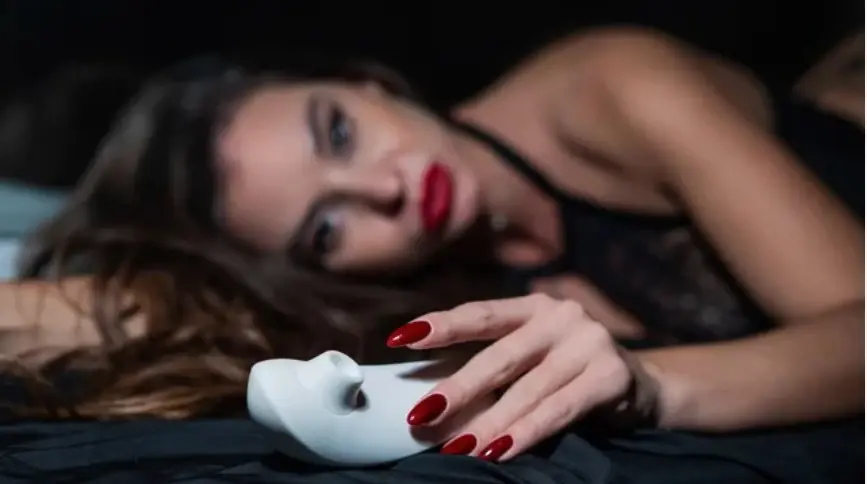 Woman lying on a bed holding a white clitoral stimulator, symbolizing relaxation and self-pleasure exploration.